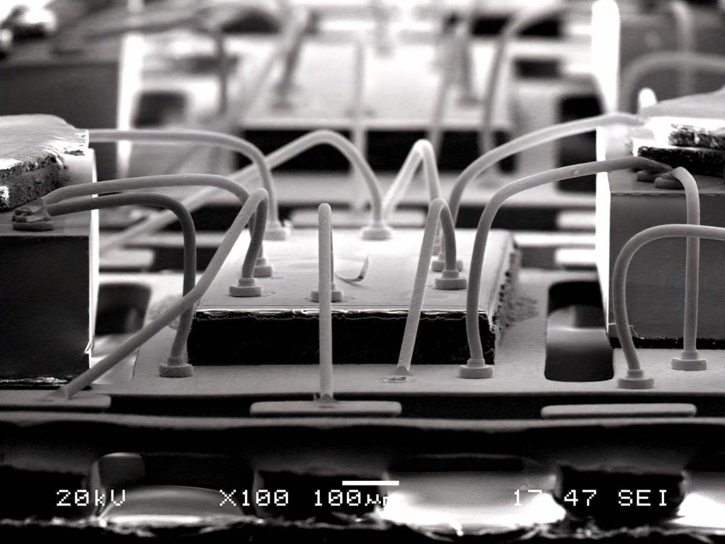 走査型電子顕微鏡(SEM)で100倍に拡大撮影されたワイヤボンディングの様子。半導体チップ上のパッドからボールボンド接合部を起点に複数のワイヤがループ状に立ち上がり、周囲のリードへ接続されている