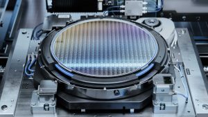 半導体製造装置のステージ上にセットされた300mmウェハのクローズアップ。表面に微細な回路パターンが格子状に形成されており、虹色の光沢を帯びている